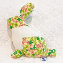 Load image into Gallery viewer, A handmade quilted fabric bunny cushion with a floral pattern in pink, green, and yellow on a white background, resting on a white quilted blanket. A small sewn label is visible on the side.
